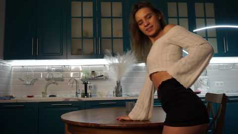 Media: Video of a slender Caucasian woman with long brown hair, wearing a white knit top and black shorts, leaning on a wooden table in a modern teal kitchen.