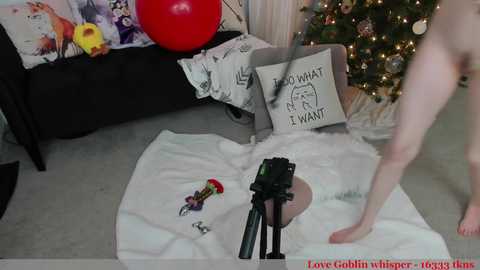 Media: A video of a nude, pale-skinned woman standing on a white bedsheet in a living room. She holds a camera, facing a decorated Christmas tree. Toys and a red ball are scattered around.