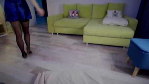 Video of a woman in a blue dress, black stockings, and black heels walking away from a bright yellow sectional sofa with white pillows in a modern living room with light wood flooring.