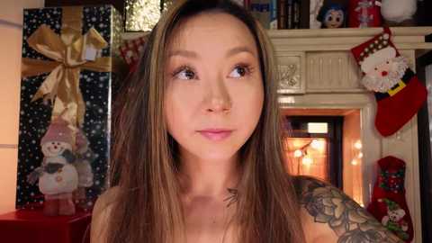 Media: Video of an Asian woman with long brown hair, tattoos, and a neutral expression in a warmly lit room with holiday decorations, including a Santa and a snowman gift wrap.