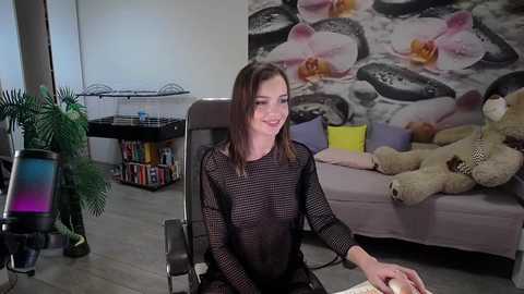 Media: Video of a young woman with straight brown hair, wearing a sheer black mesh top, sitting on a black office chair in a modern living room with plush toys and floral wallpaper.