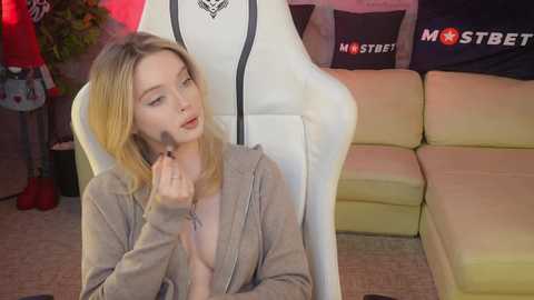 Media: Video of a young woman with fair skin and blonde hair, wearing a grey hoodie, applying makeup while seated in a white gaming chair.