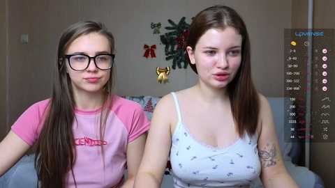 Media: Video of two young Caucasian women with straight, long brown hair, one wearing glasses and a pink shirt, the other a white camisole. They sit on a bed with a Christmas tree in the background.