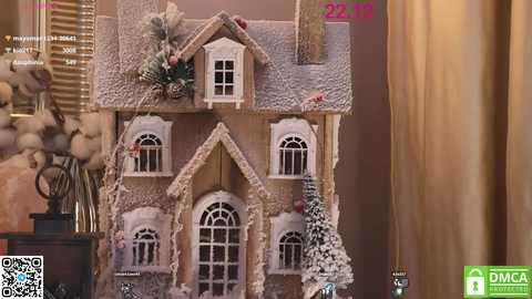 Media: A video of a detailed, miniature wooden house model with snow-covered roof, adorned with greenery, placed on a brown wooden surface.