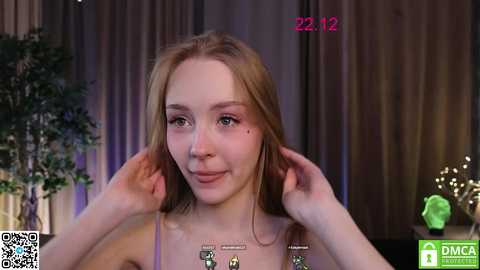 Media: Video of a young Caucasian woman with fair skin and light brown hair, smiling, adjusting her hair, in a cozy indoor setting with greenery and a camera watermark.