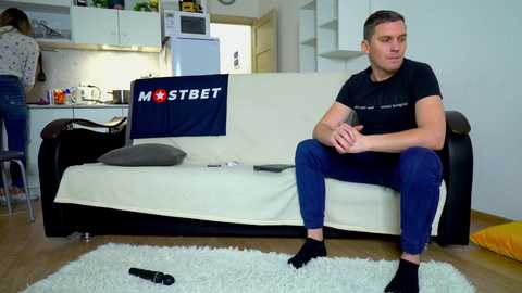 Media: Video of a young man with short hair, wearing a black t-shirt and blue jeans, sitting on a white sofa in a modern kitchen-living room.