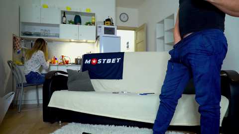 Media: Video of a modern kitchen with white cabinets and a black couch. A man in blue jeans and a black shirt is standing on the couch, while a woman in a white dress sits at a table.