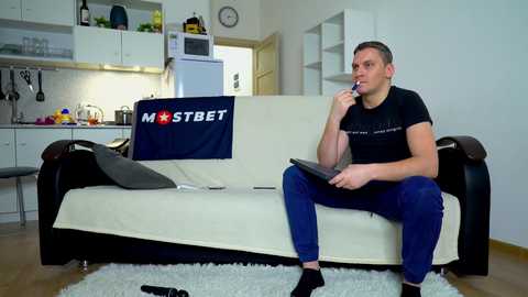 Media: Video of a muscular, clean-shaven man in a black t-shirt and blue jeans, sitting on a white couch in a modern kitchen, holding a phone to his ear, with a \"Mostbet\" flag draped on the couch.