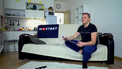 Media: Video of a man sitting on a beige sofa in a modern, minimalist kitchen-living space, wearing a black t-shirt and blue pants, holding a remote.