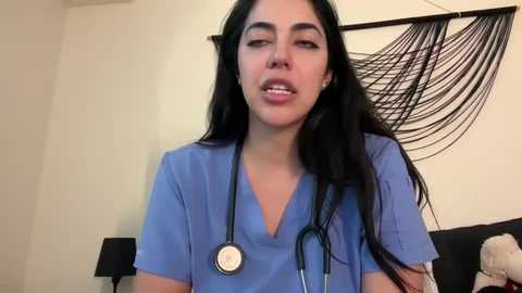 Media: Video of a young woman with long black hair, wearing blue scrubs, holding a stethoscope, standing in a bedroom with a black hammock and stuffed animals.