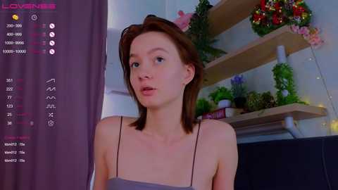 Media: Video of a fair-skinned, young woman with shoulder-length auburn hair, wearing a light gray spaghetti-strap top, sitting indoors with floral decor behind her.