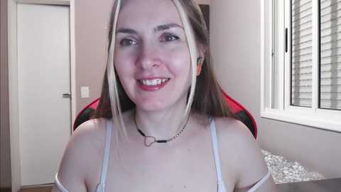 Media: A video of a young Caucasian woman with fair skin, long blonde hair, and a slight smile. She wears a white camisole and a black choker necklace. The background shows a white door and a red gaming chair.