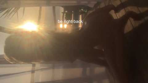 Media: A video of a person, likely a woman, lying on a bed with a sunlit window in the background, creating a warm, golden glow. The text \"be right back\" is overlaid in white font.