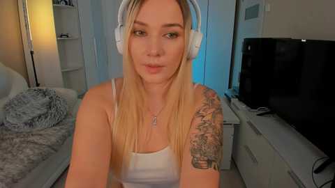 Media: A video of a blonde woman with light skin and a tattooed arm wearing a white tank top, sitting in a modern bedroom with a TV, bed, and white furniture.