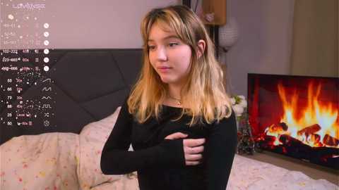 Media: A video shows a young Asian woman with shoulder-length blonde hair, wearing a black long-sleeve top, standing in a dimly lit bedroom with a lit fireplace and a bed with floral sheets.