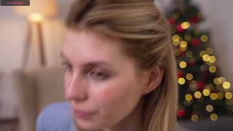 Media: A blurry video of a fair-skinned woman with blonde hair styled in a messy bun, smiling, in a warmly lit room with a Christmas tree adorned with red and gold ornaments.