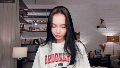 Video of a young woman with long black hair, wearing a white \"ROCKY 1988\" t-shirt, standing in a cozy, book-filled room with white brick walls, a lamp, and shelves.