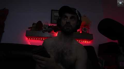 Media: Video of a bearded man in his early 30s, shirtless with chest hair, wearing headphones, seated in dimly lit room with red LED lights, plush toys, and a microphone in the background.