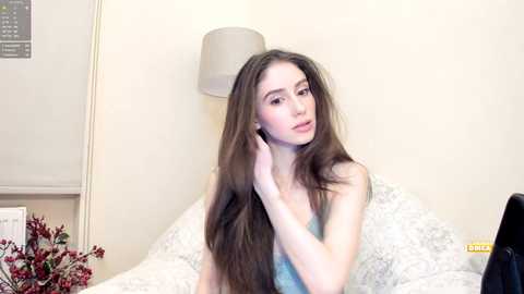 Media: Video of a young Caucasian woman with long, straight brown hair, fair skin, and a slender physique, wearing a light blue tank top, sitting on a bed with white, patterned sheets.