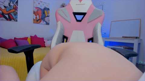 Media: Video of a person lying face-down on a yellow-striped bed with a pink and white gaming chair behind them, featuring a colorful wall art and a white desk.