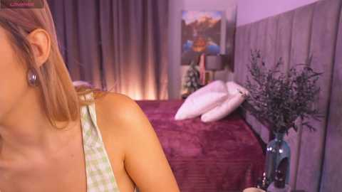 Video of a woman with light skin, wearing a green plaid tank top, standing beside a bed with a maroon bedspread and white pillows. A vase of dried leaves sits on a bedside table.