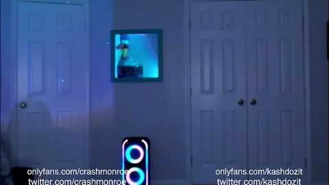 Media: Video of a dimly lit bedroom with a flat-screen TV mounted on the wall, showing a blue-tinted image. A tall, black speaker with glowing blue lights stands in front of a closed white door. The walls are painted in a soft, neutral hue.