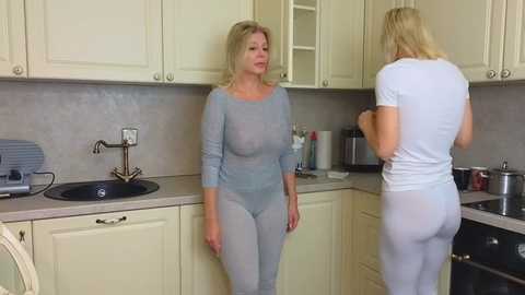 Media: Video of a blonde woman in a sheer light blue bodysuit, standing in a modern kitchen with beige cabinets, countertop appliances, and another woman in a white outfit.