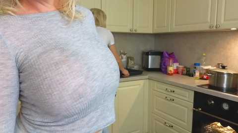 Media: Video of a woman with large breasts in a tight, light gray sweater, standing in a modern kitchen with white cabinets, a coffee maker, and a purple gift bag on the counter.