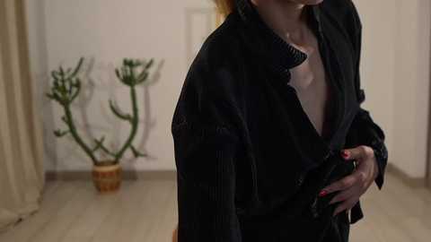 Media: A video of a fair-skinned woman with long, light brown hair, wearing a black, oversized, pinstripe coat, indoors, holding a white shirt, with green cacti in the background on a light wooden floor.