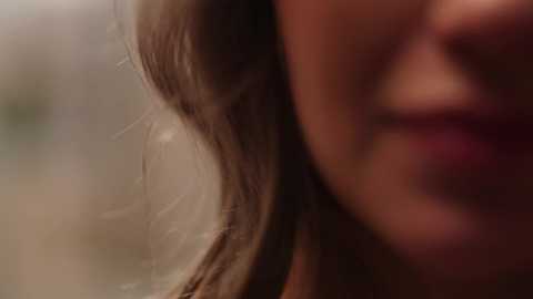 Media: Close-up video of a woman's face, focusing on her hair and partially visible lips. The background is blurred, emphasizing the texture of her light brown hair. The image has a soft, warm tone.