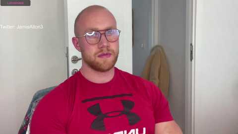 Media: Video of a bald, bearded man with glasses and a red Under Armour shirt, standing indoors next to a white door and a beige towel.