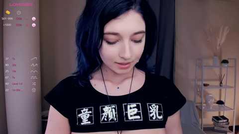 Media: A video of a young woman with long, dark blue hair, wearing a black crop top with Japanese characters and a necklace. She stands indoors with a modern, minimalist background.