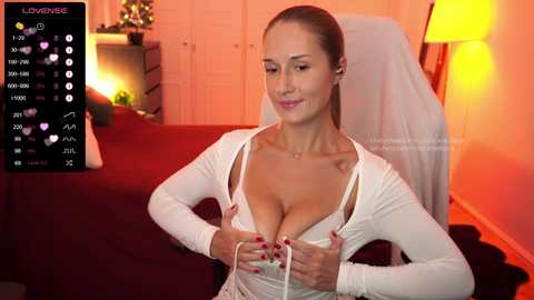 Media: Video of a fair-skinned, slender woman with medium-sized breasts, wearing a white, long-sleeve, off-shoulder top, sitting on a bed, adjusting a white bra, in a warmly-lit bedroom.