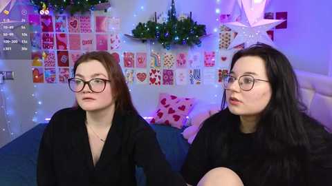 Media: Video of two women with glasses, one in a black dress, one in a black sweater, seated on a blue couch in a festive, decorated room with heart-themed posters and twinkling lights.