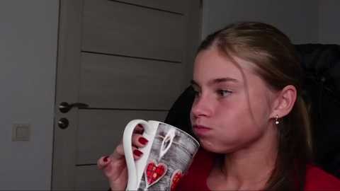 Media: A video of a young woman with light skin and brown hair, wearing a red top, drinking from a white mug with a red and black pattern, sitting in a black chair against a white wall with a closed door.