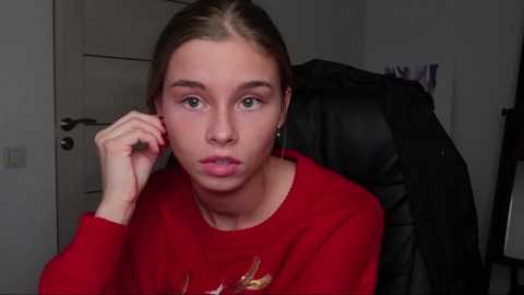 Media: Video of a young Caucasian woman with fair skin and brown hair in a red sweater, sitting in a chair, looking pensive, with a black jacket hanging behind her.