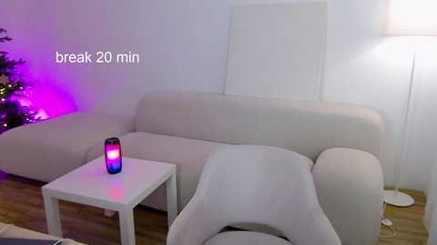 Media: Video of a minimalist living room featuring a white sectional sofa, a small white side table, and a glowing purple and blue candle. The background includes a partially visible Christmas tree with multicolored lights.