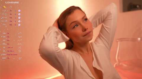 A video of a fair-skinned woman with light brown hair, wearing a white cardigan, looking serene with hands behind her head, in a warmly lit room.