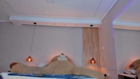 Media: Video of a modern, minimalist bedroom with beige walls, a plush bed covered in light brown and blue bedding, three pendant lamps with red shades, and a vertical wooden panel.