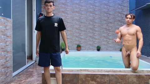 Media: Video of two men in a modern, outdoor pool area; one is shirtless, muscular, and circumcised, the other is dressed in a black t-shirt and dark shorts.