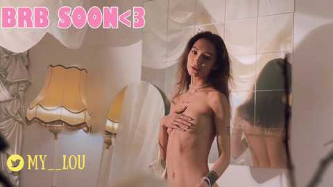 Media: A video of a topless, slender, East Asian woman with long dark hair, standing in front of a mirror in a luxurious bathroom with beige tiles and a gold lamp.