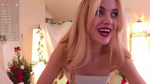 Media: Video of a smiling, fair-skinned woman with long blonde hair, wearing a strapless white dress, standing in a warmly lit, festive room with Christmas decorations.