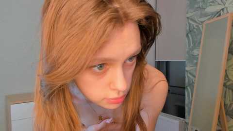 Media: A close-up video of a fair-skinned, young girl with long, straight, auburn hair, wearing a light pink top, looking downward with a thoughtful expression. Background shows a white door and green wallpaper.