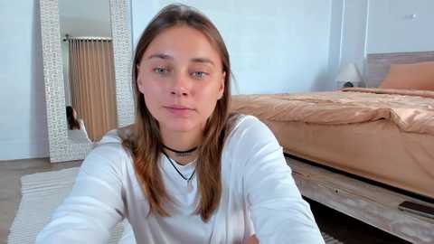 Media: Video of a young woman with straight brown hair, wearing a white long-sleeve shirt, sitting on the floor in a modern bedroom with a beige bed, wooden floor, and large mirror.