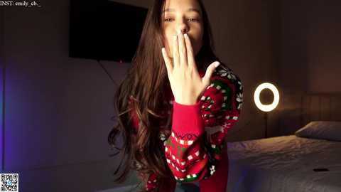 Media: Video of a young woman with long brown hair, wearing a red Christmas sweater, covering her mouth with her hand, standing in a dimly lit bedroom with a TV and round lamp.