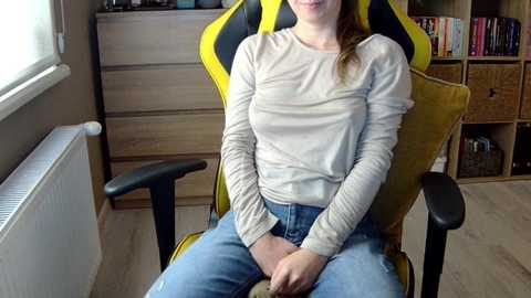 Media: Video of a woman in a beige long-sleeve shirt and blue jeans sitting in a yellow and black gaming chair in a cozy room with wooden furniture, books, and a window.