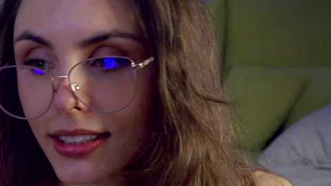 Video of a young woman with long, wavy brown hair, wearing round glasses and light makeup. She has a fair complexion and is smiling slightly. Background shows a green pillow and blurred bedroom details.