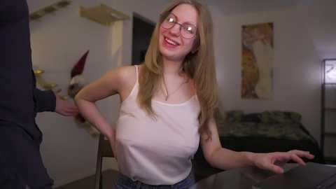 Media: Video of a smiling, glasses-wearing, light-skinned woman with long blonde hair, wearing a white tank top, sitting on a bed in a dimly-lit bedroom.