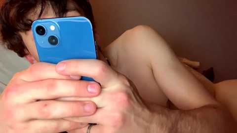 Media: Video of a nude person with fair skin and short brown hair, holding a blue smartphone to take a selfie, lying on a bed with white sheets.