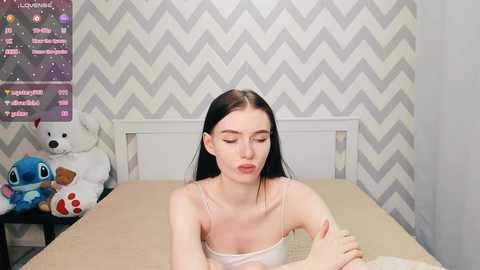 Media: Video of a pale-skinned, long-haired woman in a white tank top sitting on a bed with beige bedding, surrounded by chevron-patterned wallpaper, plush toys, and a digital clock.